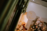A decorative setup featuring an arrangement of various flowers in soft pastel hues. There is a circular sign with gold lettering against a neutral background. The setting conveys an elegant and sophisticated atmosphere, possibly for a wedding or special event. The lighting is warm, adding a cozy ambiance.