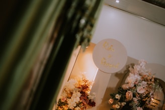 A decorative setup featuring an arrangement of various flowers in soft pastel hues. There is a circular sign with gold lettering against a neutral background. The setting conveys an elegant and sophisticated atmosphere, possibly for a wedding or special event. The lighting is warm, adding a cozy ambiance.