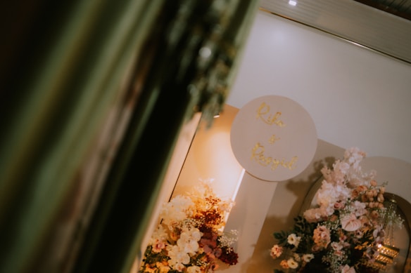 A decorative setup featuring an arrangement of various flowers in soft pastel hues. There is a circular sign with gold lettering against a neutral background. The setting conveys an elegant and sophisticated atmosphere, possibly for a wedding or special event. The lighting is warm, adding a cozy ambiance.