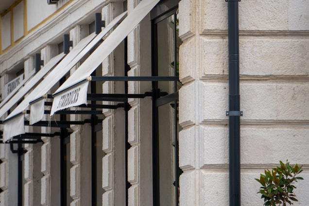A row of white building awnings are attached to a stone facade. The wall is textured with rectangular patterns and there is a prominent black drainpipe running vertically. Some foliage is visible in the corner, suggesting nearby plants or landscaping.