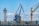 A large industrial crane alongside a tower structure, with a backdrop of trees and hills under a partly cloudy sky. The crane is blue with a distinctive arm extended outward. Various industrial items are located near the base, and there is another crane in the background.