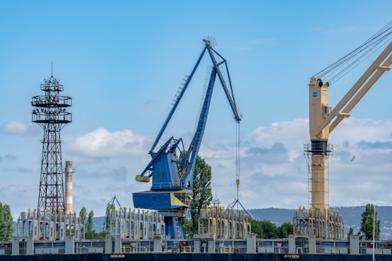A large industrial crane alongside a tower structure, with a backdrop of trees and hills under a partly cloudy sky. The crane is blue with a distinctive arm extended outward. Various industrial items are located near the base, and there is another crane in the background.