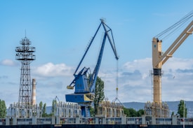 A large industrial crane alongside a tower structure, with a backdrop of trees and hills under a partly cloudy sky. The crane is blue with a distinctive arm extended outward. Various industrial items are located near the base, and there is another crane in the background.