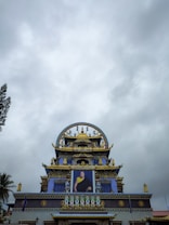 An ornate Buddhist temple featuring intricate architectural details with vibrant colors and gilded elements. A large portrait is prominently displayed on the facade, surrounded by intricate decorations and symbols. The sky is overcast with a cloudy backdrop.
