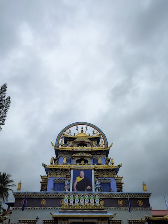 An ornate Buddhist temple featuring intricate architectural details with vibrant colors and gilded elements. A large portrait is prominently displayed on the facade, surrounded by intricate decorations and symbols. The sky is overcast with a cloudy backdrop.