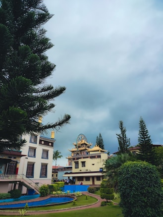 A serene landscape featuring a prominent temple structure with intricate architecture surrounded by lush greenery. There is a large tree in the foreground, and smaller shrubs and a neatly maintained garden path are visible. The sky is overcast, giving the scene a tranquil and peaceful ambiance.