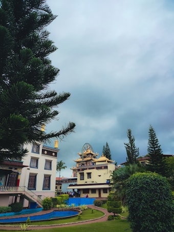 A serene landscape featuring a prominent temple structure with intricate architecture surrounded by lush greenery. There is a large tree in the foreground, and smaller shrubs and a neatly maintained garden path are visible. The sky is overcast, giving the scene a tranquil and peaceful ambiance.