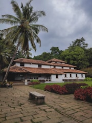 A traditional white building with a clay-tiled roof is surrounded by lush green vegetation. Tall palm trees tower over the structure, and vibrant red shrubs line the walkway leading to the entrance. A stone bench is situated on a paved stone path.