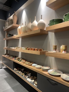 A series of wooden shelves displaying various ceramic items such as vases, mugs, bowls, and plates. Large, elegantly shaped vases stand prominently on the higher shelf, while smaller mugs and dishes occupy the lower shelves. The shelves are lit by overhead lighting, enhancing the earthy tones of the ceramics.