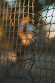 A close-up view of a weathered net with visible wear and tear. The net is woven with coarse material, and some sections are frayed and patched with black string. The background is out of focus, with warm sunlight creating a serene ambiance.