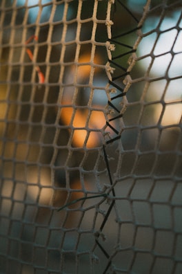 A close-up view of a weathered net with visible wear and tear. The net is woven with coarse material, and some sections are frayed and patched with black string. The background is out of focus, with warm sunlight creating a serene ambiance.