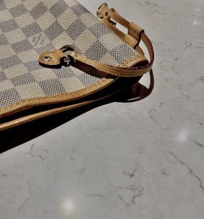 A close-up view of a designer checkered pattern handbag with beige and dark gray squares. The bag features a leather strap with metal hardware lying on a smooth, light-colored marble surface.