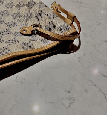 A close-up view of a designer checkered pattern handbag with beige and dark gray squares. The bag features a leather strap with metal hardware lying on a smooth, light-colored marble surface.