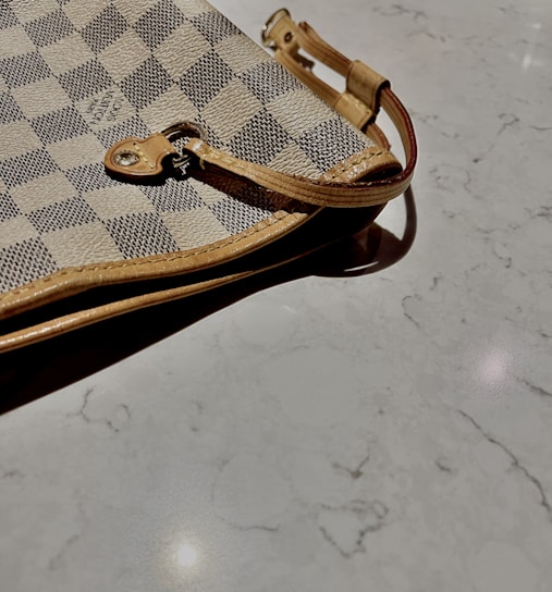 A close-up view of a designer checkered pattern handbag with beige and dark gray squares. The bag features a leather strap with metal hardware lying on a smooth, light-colored marble surface.
