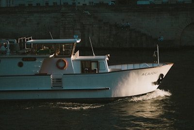 A white boat named Acajou is cruising through the water. The vessel has a cabin with large windows and a lifebuoy attached to its side. The boat is illuminated by low, warm sunlight, casting soft shadows on its surface. In the background, an old stone wall with some steps and seated people can be seen, partially in shadow.