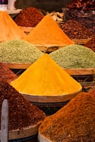 Colorful piles of spices arranged in conical shapes are displayed in a market. The spices vary in colors including vibrant yellows, oranges, reds, and greens, each presented in its own container.