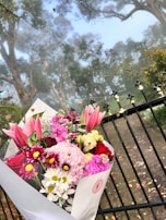 A beautiful bouquet of assorted flowers, including lilies, chrysanthemums, daisies, and carnations in vibrant colors such as pink, red, white, and burgundy, is set against the backdrop of a misty forest. The bouquet is wrapped in white paper and positioned in front of a black metal fence with heart-shaped decorative elements.