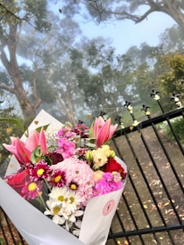 A beautiful bouquet of assorted flowers, including lilies, chrysanthemums, daisies, and carnations in vibrant colors such as pink, red, white, and burgundy, is set against the backdrop of a misty forest. The bouquet is wrapped in white paper and positioned in front of a black metal fence with heart-shaped decorative elements.