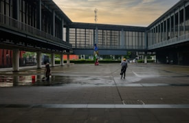 An open, spacious courtyard is flanked by modern, glass-walled buildings with overhanging walkways. Two workers, one with a mop and a red bucket and the other with a hose, are cleaning the wet pavement. The sky is lightly clouded, indicating early morning or late afternoon.