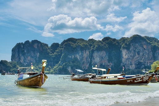 Several traditional longtail boats are floating on clear turquoise water near a picturesque coastline with lush, towering limestone cliffs in the background under a partly cloudy sky.
