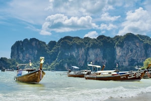 Several traditional longtail boats are floating on clear turquoise water near a picturesque coastline with lush, towering limestone cliffs in the background under a partly cloudy sky.
