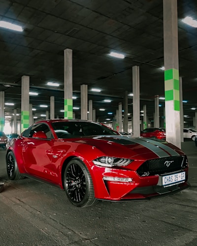 A red sports car with black wheels is parked in an indoor parking lot with concrete pillars and green markings. The car has a license plate reading CHAS 25 GP and is adorned with black racing stripes on the hood. The lighting is artificial, from overhead fluorescent lights.