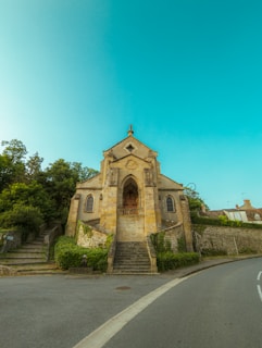 An old stone church with arched doorways and small windows sits at the top of a staircase. The building is flanked by green shrubs and trees, with a curved road leading around it. The sky is clear and bright, creating a tranquil atmosphere.