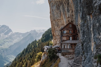 A rustic wooden building is perched against a steep cliff, with a scenic view of mountainous terrain and dense forest below. The structure appears to be integrated into the natural rock formation, surrounded by greenery and overlooking distant mountain peaks.