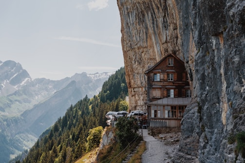 A rustic wooden building is perched against a steep cliff, with a scenic view of mountainous terrain and dense forest below. The structure appears to be integrated into the natural rock formation, surrounded by greenery and overlooking distant mountain peaks.
