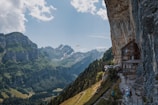 A picturesque scene of a mountain landscape with a chalet built into the side of a rocky cliff. The distant backdrop is filled with towering, snow-capped mountain peaks and lush greenery. A paraglider sails through the clear blue sky. In the foreground, several people are walking along a narrow path near the chalet, admiring the view.