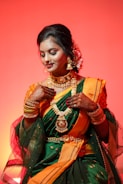 A woman dressed in an elegant traditional attire with intricate henna designs on her hands. She is adorned with elaborate gold jewelry, including necklaces, bangles, and earrings. Her sari is green with orange and gold accents, and she has flowers in her hair, set against a warm orange background.
