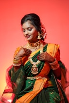 A woman dressed in an elegant traditional attire with intricate henna designs on her hands. She is adorned with elaborate gold jewelry, including necklaces, bangles, and earrings. Her sari is green with orange and gold accents, and she has flowers in her hair, set against a warm orange background.