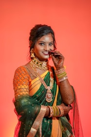 A woman dressed in a traditional Indian outfit, featuring a vibrant orange and green saree with elaborate gold jewelry. Her hands are adorned with intricate henna designs, and she wears several colorful bangles. Her hair is styled neatly, and she appears to be in a festive mood against a rich, warm orange background.