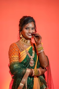 A woman dressed in a traditional Indian outfit, featuring a vibrant orange and green saree with elaborate gold jewelry. Her hands are adorned with intricate henna designs, and she wears several colorful bangles. Her hair is styled neatly, and she appears to be in a festive mood against a rich, warm orange background.