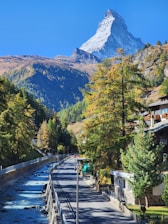 A scenic mountain landscape features a prominent, snow-capped peak in the background beneath a clear, blue sky. In the foreground, a road runs alongside a flowing river, lined with trees bearing green and autumnal yellow foliage. Residential buildings are visible on the right side of the image, partially obscured by the tall trees.