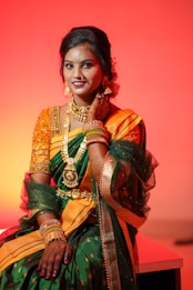 A woman dressed in a traditional Indian sari adorned with elaborate gold jewelry, including a necklace and earrings. The sari is vibrant with shades of green and orange, and she has intricate mehndi designs on her hands. The background is a reddish-orange hue, creating a warm and celebratory atmosphere.