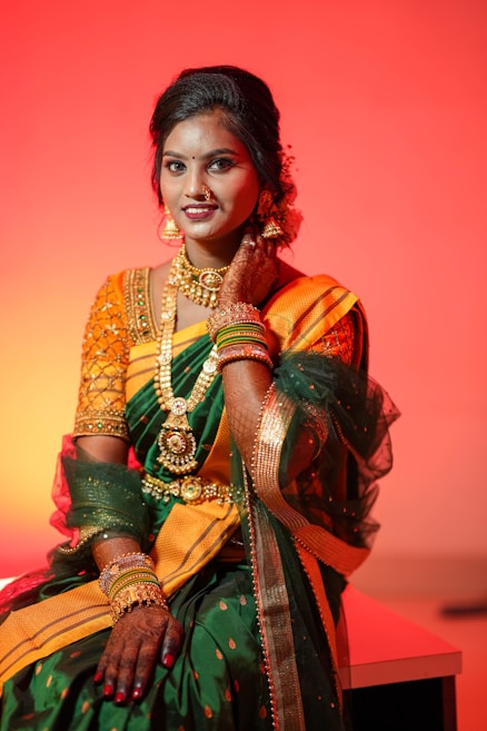 A woman dressed in a traditional Indian sari adorned with elaborate gold jewelry, including a necklace and earrings. The sari is vibrant with shades of green and orange, and she has intricate mehndi designs on her hands. The background is a reddish-orange hue, creating a warm and celebratory atmosphere.