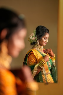 A woman wearing traditional Indian attire, adorned with intricate jewelry and floral hair accessories, is looking downwards with a serene expression. Her outfit features rich green and yellow fabrics with elaborate patterns. The scene is reflected in a mirror, adding depth to the composition.