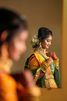 A woman wearing traditional Indian attire, adorned with intricate jewelry and floral hair accessories, is looking downwards with a serene expression. Her outfit features rich green and yellow fabrics with elaborate patterns. The scene is reflected in a mirror, adding depth to the composition.