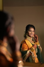 A woman wearing a traditional outfit with a yellow and green saree adorned with intricate embroidery is smiling and adjusting her earrings. She has henna on her hands and is wearing gold jewelry including bracelets, necklaces, and earrings. The setting appears to be indoors, reflected in a mirror with a soft focus on the background.