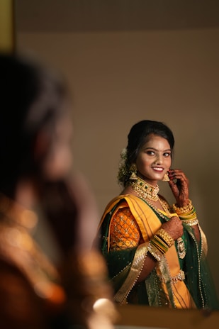 A woman wearing a traditional outfit with a yellow and green saree adorned with intricate embroidery is smiling and adjusting her earrings. She has henna on her hands and is wearing gold jewelry including bracelets, necklaces, and earrings. The setting appears to be indoors, reflected in a mirror with a soft focus on the background.