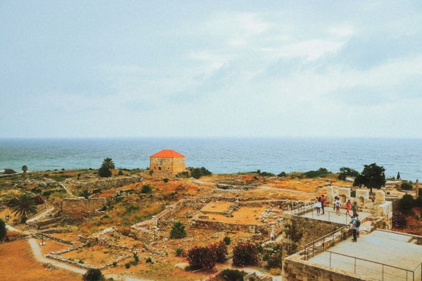 A coastal landscape featuring ancient ruins with stone walls and pathways. A house with a red roof stands out against the blue sea in the background. Several people are visible exploring the site, adding a sense of scale to the expansive view. The area is dotted with green shrubs and trees, enhancing the historical atmosphere.