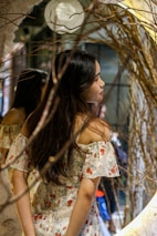 A model wearing a flowing, dark floral dress standing amidst twisted branches and moss.