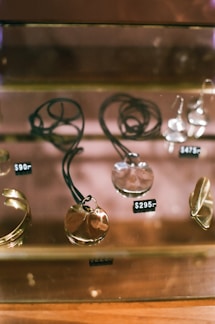 A display case containing jewelry pieces including necklaces and rings. The items have price tags such as $90, $295, and $475, indicating value. The jewelry pieces are arranged neatly on a reflective surface, enhancing their metallic sheen.