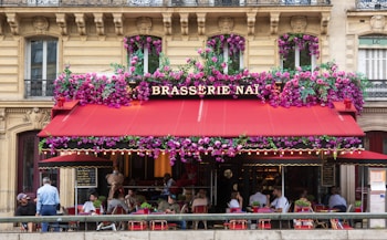 A charming café with a bright red awning, adorned with an abundance of pink and purple flowers, is situated on a traditional European street. People are seated at tables on the sidewalk, enjoying food and drinks. The architecture of the building features intricate stonework with tall windows, giving it an elegant, historic appearance.