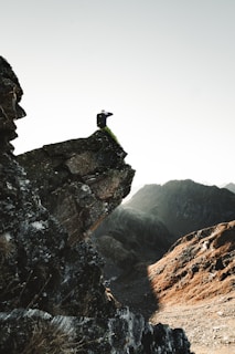A person sits on the edge of a steep rock formation overlooking a rugged mountain landscape. The scene captures a sense of adventure and vastness, with the sun casting shadows over the rocky terrain.