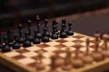 A wooden chessboard with a full set of black pieces set up in the foreground. The pieces are intricately carved, featuring details on the knights, bishops, queens, kings, and pawns. The chessboard has alternating light and dark squares, creating a classic checkered pattern. The focus is on the black side, with blurred brown and beige pieces visible in the background, suggesting an ongoing or upcoming game.