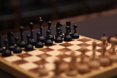 A wooden chessboard with a full set of black pieces set up in the foreground. The pieces are intricately carved, featuring details on the knights, bishops, queens, kings, and pawns. The chessboard has alternating light and dark squares, creating a classic checkered pattern. The focus is on the black side, with blurred brown and beige pieces visible in the background, suggesting an ongoing or upcoming game.