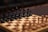 A wooden chessboard with a full set of black pieces set up in the foreground. The pieces are intricately carved, featuring details on the knights, bishops, queens, kings, and pawns. The chessboard has alternating light and dark squares, creating a classic checkered pattern. The focus is on the black side, with blurred brown and beige pieces visible in the background, suggesting an ongoing or upcoming game.