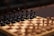 A wooden chessboard with a full set of black pieces set up in the foreground. The pieces are intricately carved, featuring details on the knights, bishops, queens, kings, and pawns. The chessboard has alternating light and dark squares, creating a classic checkered pattern. The focus is on the black side, with blurred brown and beige pieces visible in the background, suggesting an ongoing or upcoming game.